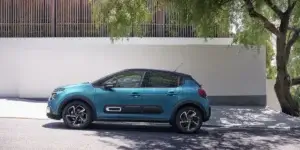 Side view of a Citroën C3 car in motion on a road. The car is a compact hatchback with a distinctive two-tone paint job, featuring a white body and a contrasting black roof. The vehicle is driving in an urban environment, with blurred buildings and greenery in the background.