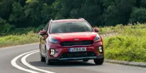 A red Kia Niro driving on a winding road surrounded by trees. The car is facing the camera and has a sleek, modern design. The license plate on the car reads "LF18 KJG.