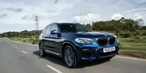 A blue BMW X3 SUV driving on a road with a clear sky and a power plant in the background. The car has a sporty design with large alloy wheels and a prominent grille.