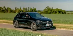 A black Mercedes-Benz GLA SUV driving down a country road with a field of green grass on either side.