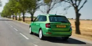 A green Skoda Fabia Combi estate car driving on a road with trees and a field in the background. The car is in motion, and the rear of the car is visible, showing the taillights, rear bumper, and license plate.