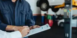 A photo of two mechanics wearing a blue uniform and a clipboard. The mechanic's face is not visible. The background is blurred and shows a garage workshop.