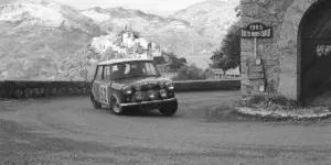 A black and white photo of a classic Mini Cooper rally car driving on a winding mountain road. The car is surrounded by lush greenery and a stone building is visible in the background.