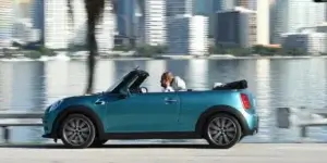 A blue MINI Cooper Convertible driving along a coastal road with a cityscape in the background. The top is down, and the driver is wearing sunglasses and a hat.