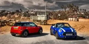 Two MINI Coopers parked side-by-side on a beach. The car on the left is red, and the car on the right is blue. The ocean and a beach house are visible in the background.