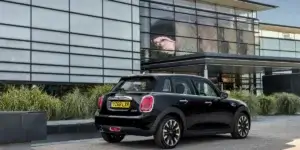 A black MINI Cooper 5-door hatchback parked in front of a modern building. The car is facing away from the camera and has a sleek, sporty design.