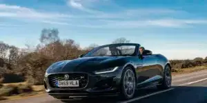 A black Jaguar F-Type convertible driving on a winding road surrounded by trees. The car's top is down, and two people are visible in the front seats.