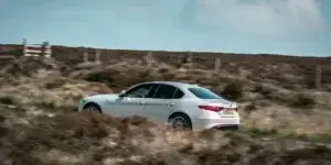 A white Alfa Romeo Giulia driving on a dirt road surrounded by rolling hills and a fence. The car is viewed from the rear and features a sporty design with alloy wheels.