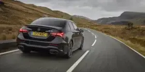 A black Mercedes-Benz A-Class AMG sedan driving on a winding road surrounded by mountains. The car is viewed from the rear and features a sporty design with dual exhaust pipes and a black license plate that reads "KY71 HBG.