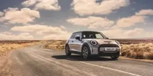 The image shows a silver MINI Cooper 3-door hatchback driving on a winding road in a rural landscape. The sky is blue with white clouds, and the road is surrounded by green fields.