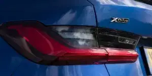 A close-up of the rear taillight of a blue BMW 3 Series car. The taillight has a sleek and modern design with LED elements and the "xDrive" badge indicating all-wheel drive.