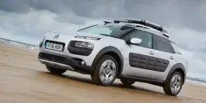 The image shows a silver Citroën C4 Cactus driving on a beach. The car has a roof rack with a surfboard attached to it. The tide is low and the sand is wet. The car has a rugged appearance with black plastic cladding around the wheel arches and side skirts. The license plate on the car reads "KT16 HON.