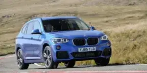 A blue BMW X1 SUV driving on a winding road with a scenic landscape in the background. The car has a sporty design with large alloy wheels and a prominent grille.