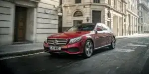 A red Mercedes-Benz E-Class sedan is driving down a narrow street in a city. The car has alloy wheels and is surrounded by buildings.