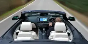 A photo from inside a BMW 6 Series Convertible. The driver is visible, wearing a black shirt and holding the steering wheel. The top is down and the white leather seats are visible. The dashboard features a navigation screen and the car is driving on a winding road.