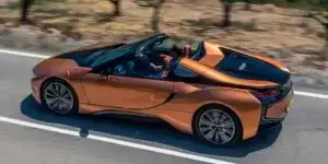 A bronze-colored BMW i8 Roadster driving down a winding road with a scenic landscape in the background. The top is down.