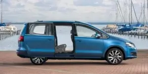 The image shows a blue Volkswagen Sharan parked next to a body of water. The sliding door on the passenger side is open, revealing the interior of the car. There are several sailboats visible in the background.