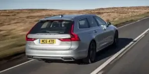 A silver Volvo V90 R-Design wagon driving on a rural road. The car is viewed from the rear and features a sporty design with dual exhaust pipes and a black license plate that reads "MCG 470.