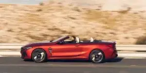 A red BMW M8 Convertible driving down a highway with a desert landscape in the background. The top is down and the driver is visible.