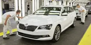 The image you uploaded shows a white car on a production line with two workers inspecting it. The car appears to be a Škoda Superb, based on its distinctive grille and design features. The setting seems to be an automotive manufacturing or quality control area, where cars are inspected for any imperfections before they are sent out for delivery. The workers are dressed in white uniforms, which is typical in such environments to maintain cleanliness and reduce contamination.