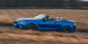 A blue BMW M4 Convertible driving on a dirt road with a field of grass in the background. The top is down.