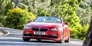 A red BMW 6 Series Convertible driving on a winding road with lush green trees on either side. The top is down and two people are visible in the car.