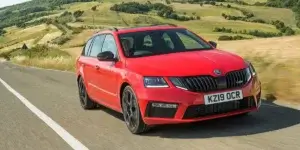 A red Skoda Octavia RS estate car driving on a winding road with a scenic landscape in the background. The car has a sporty design with large alloy wheels and a prominent grille. The license plate on the car is KZ19 OCR.