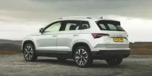 A white Skoda Karoq SUV parked on a dirt road with a scenic landscape in the background. The rear of the car is visible, showing the taillights, roof rails, and alloy wheels.