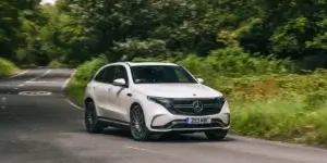 The car in the image is a white Mercedes-Benz EQC. It is an electric SUV that is driving down a winding road in a rural area. The car has a modern and sleek design, with large wheels and a black grille. The road is surrounded by trees and greenery.