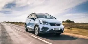 The image shows a blue Honda Jazz Crosstar driving on a country road. The car is facing the camera and has a sleek, modern design. 