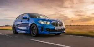 The image shows a blue BMW 1 Series driving on a road at sunset. The car is facing the camera and has a sporty design with large air intakes and aggressive styling. The sky is orange and pink, and the sun is setting behind the car. The car is driving at a high speed, and the road is straight and empty.