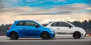 The image shows two Abarth 595 cars parked side-by-side on a racetrack. The car on the left is blue, and the car on the right is white. Both cars have black wheels and black accents. The background of the image shows a clear blue sky with mountains in the distance.