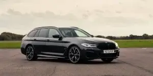 The image shows a black BMW 5 Series Touring. The car is parked on a tarmac road with a runway in the background. The car has a sleek and sporty design, with a muscular front end and large alloy wheels. The rear of the car features a sloping roofline and a large tailgate.