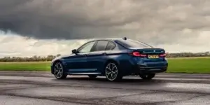 The image shows a blue BMW 5 Series driving on a wet road. The car is seen from the rear and features sporty styling with large alloy wheels and a prominent rear diffuser. The sky is cloudy and dramatic, suggesting stormy weather.