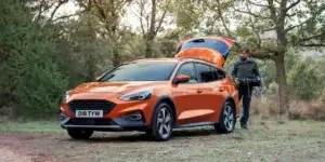 A man with a backpack is standing next to an orange Ford Focus Active wagon. The car's tailgate is open, and the man is reaching inside. The car is parked in a forest setting.