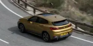 A gold BMW X2 SUV driving on a winding road with a scenic landscape in the background. The car is in motion, and the rear of the car is visible, showing the taillights, rear bumper, and license plate.