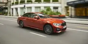 A red BMW 3 Series sedan is driving down a busy city street. The car has alloy wheels and is moving at a high speed.