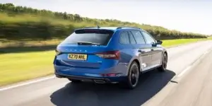 A blue Skoda Superb Combi estate car driving on a road with a field and a clear sky in the background. The car is in motion, and the rear of the car is visible, showing the taillights, rear bumper, and license plate.