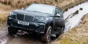 A black BMW X7 SUV driving through a muddy off-road track. The car has all-terrain tires and is equipped with features for off-road driving.