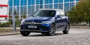 The car in the image is a blue Mercedes-Benz GLC. It appears to be driving on a city street. The car has a sporty design with a large front grille and headlights. It is also equipped with a sunroof.