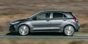 A grey Kia Rio hatchback car is driving on a road with a field in the background. The car has alloy wheels and is moving at a moderate speed.