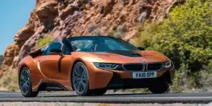 A bronze-colored BMW i8 Roadster driving on a winding coastal road with a scenic backdrop. The top is down and the driver is visible.