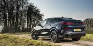A blue BMW X6 SUV parked on a dirt road surrounded by trees. The car has a sporty design with large alloy wheels and a sloping roofline.