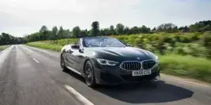 A dark green BMW 8 Series Convertible driving on a winding road surrounded by trees. The car's top is down, and it is moving at a high speed.