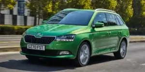 A green Skoda Fabia Combi estate car driving on a road with trees and buildings in the background. The car has alloy wheels and a modern design.