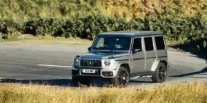 A silver Mercedes-Benz G-Class SUV driving on a winding road with a rural landscape in the background.