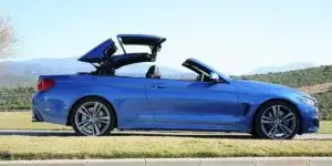 A blue BMW 4 Series Convertible with the roof partially open. The car is parked on a grassy hill with a mountain view in the background.