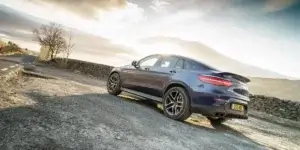 A blue Mercedes-Benz GLC Coupé parked on a gravel road with a scenic mountain backdrop. The car has black alloy wheels and a sporty rear design.