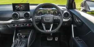 The image shows the interior of an Audi Q2. The interior is mostly black with red accents. The car has a digital instrument cluster and a large central touchscreen. The seats are sporty and supportive, with a black and gray fabric pattern. The steering wheel is wrapped in leather and has red stitching.