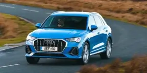 The image shows a blue Audi Q3 Sportback driving on a winding road surrounded by mountains. The car is facing the camera and has a sleek, modern design. The license plate on the car reads "AEF 150."